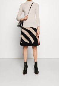 Donna che indossa un maglione a coste beige, una gonna astratta nera e beige, stivali neri alla caviglia e porta una borsa a tracolla nera.
