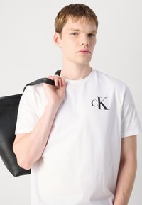 Bijela pamučna majica s crnim 'cK' logom na prsima, koju drži osoba, uz crni kožni remen torbe.