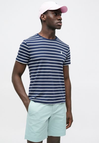 Μπλε ναυτικό και άσπρο ριγέ κοντομάνικο μπλουζάκι με στρογγυλή λαιμόκοψη, συνδυασμένο με σορτς σε μέντα πράσινο χρώμα. Μια ροζ καπέλο συμπληρώνει την εμφάνιση.