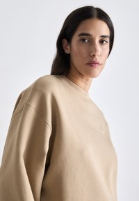 Sudadera beige con cuello redondo y hombros caídos, hecha de una suave tela texturizada. Pequeño detalle del logo en el pecho.