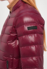 Bordo puffer jakna s horizontalno quiltiranim dijelovima, sjajnom završnom obradom i džepom na patentni zatvarač. Ima crni logo uvezen na rukavu.