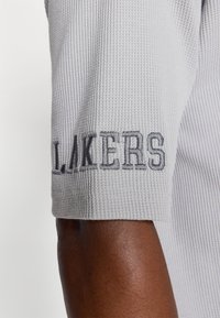 Šedá textura látková košile s krátkým rukávem, na spodním lemu vyšitý nápis "LAKERS" tmavě šedou barvou. Detailní pohled na rukáv a texturu.