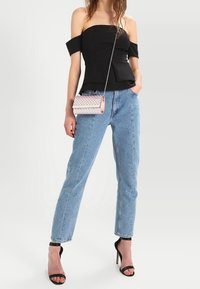 Femme portant un haut noir à épaules dénudées, un jean bleu clair, des talons hauts noirs, et portant un petit sac bandoulière rose.