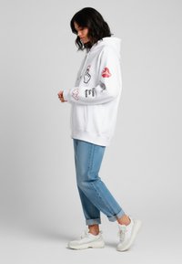 Felpa bianca con stampe grafiche, tra cui cuori e labbra, abbinata a jeans in denim blu chiaro e sneakers bianche. Look casual e vestibilità rilassata.