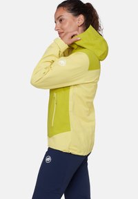 Η γυναίκα ρυθμίζει τη κουκούλα ενός διχρωμίου κίτρινου μπουφάν, φοράει σκούρο μπλε παντελόνι με λογότυπο ενός λευκού μαμούθ και στα δύο ρούχα.