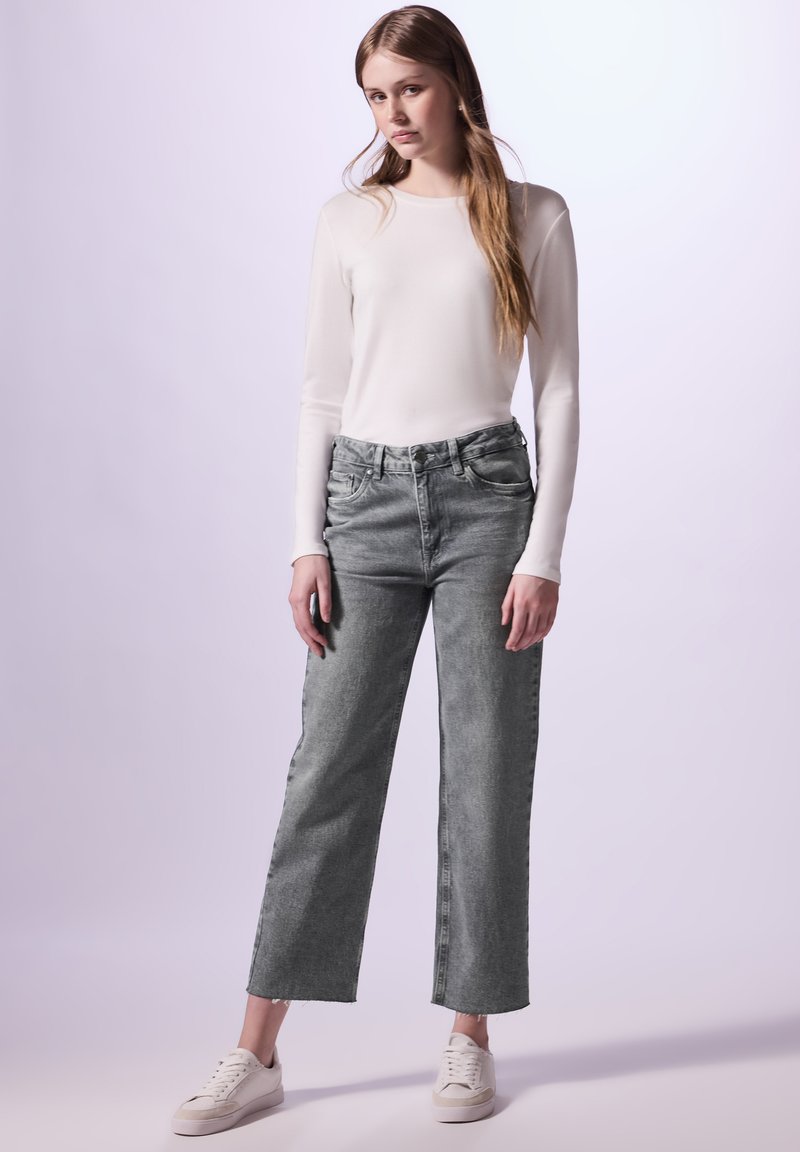 Jeans gris taille haute avec une jambe droite, ourlet effiloché, associés à un haut à manches longues blanc et des baskets blanches. Texture du tissu lisse.