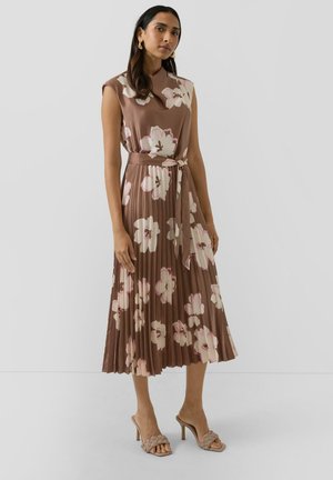 Frau trägt ein ärmelloses, braunes Midikleid mit großen weißen und pinken Blumenmustern, einem gebundenen Taillengürtel und beigefarbenen Sandaletten mit Absatz.