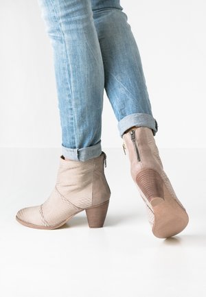 Person trägt hellblaue, hochgekrempelte Jeans und beige, absatztragende Stiefeletten mit Reißverschlüssen an den Seiten, steht auf einem weißen Boden.