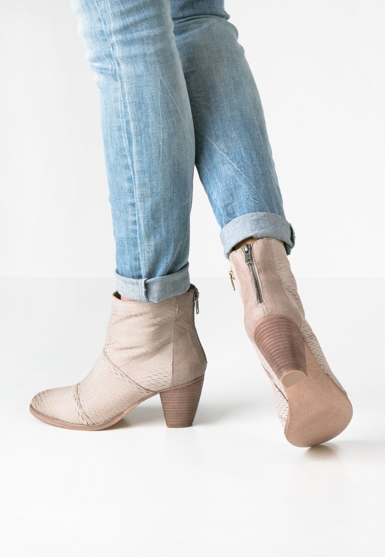 Personne portant un jean bleu clair retroussé et des bottines beiges à talons avec fermetures éclair sur les côtés, debout sur un sol blanc.