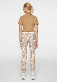 Brun polo-skjorte, mønstrede beige og hvite bukser, hvit belte og hvite sneakers med svarte detaljer. Hånd iført hansker er synlig.