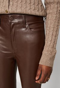 Pantalones de cuero marrón con una textura suave, que cuentan con bolsillos frontales y un cierre de botón, combinados con un suéter de punto de color beige claro.