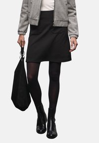 Šedá bomber bunda, černá texturovaná sukně, černé punčocháče a kotníkové boty. Modelka drží černou tašku, která doplňuje vrstvený outfit.