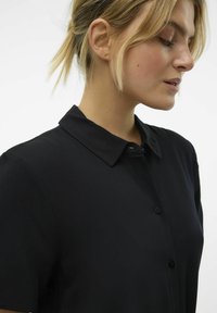 Chemise noire à bouton, avec un col. Fabriquée en tissu lisse, elle possède des manches courtes et un léger éclat.