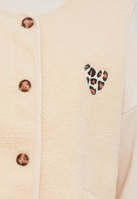 Απαλό ροζ γιλέκο από φλίς με καφέ κουμπιά, διακοσμημένο με μπαλώμα-καρδιά σε σχέδιο λεοπάρδαλη στην αριστερή πλευρά. Μαλακή υφή με ζεστή εμφάνιση.