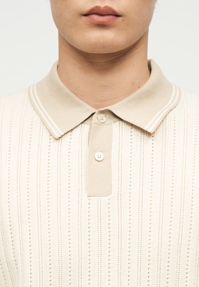 Jeune personne portant un polo en tricot beige avec un motif côtelé et deux boutons sur la patte de boutonnage, sur un fond clair.
