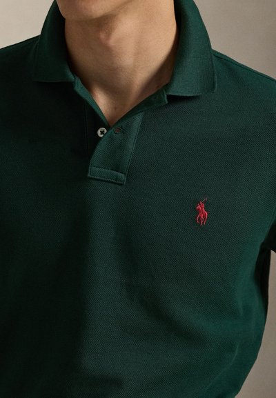 Mand iført mørkegrøn polo shirt med rødt broderet hest og rytter logo på venstre bryst, kraven delvist åbnet.