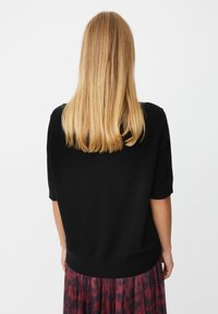 Blond kvinde med glat hår iført en sort kortærmet sweater og en rød mønstret nederdel, vist fra ryggen mod en hvid baggrund.