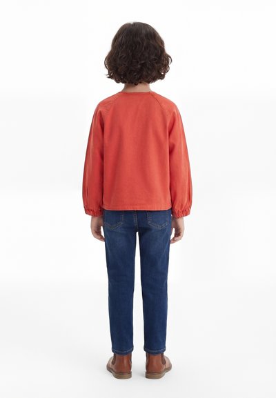 Enfant aux cheveux bouclés et foncés portant une chemise orange à manches longues, un jean bleu et des bottines brunes, debout de dos sur un fond blanc.