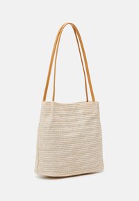 Beżowa pleciona torba na ramię z dwoma długimi, brązowymi skórzanymi paskami, stojąca prosto na tle czystego białego koloru.