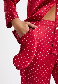 Conjunto de pijama de satén rojo con un patrón de corazones blancos, con un bolsillo que lleva una máscara para los ojos en forma de corazón a juego. Textura suave y detalles de botones.