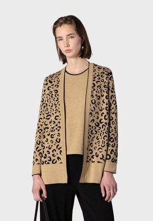 Jeune femme portant un cardigan beige à imprimé léopard par-dessus un haut assorti et un pantalon noir, tenant un sac à main noir sur un fond uni.