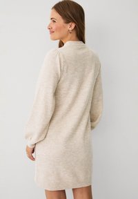 Kvinde med lys brunt hår iført en beige langærmet strikket kjole, står sidelæns med et let smil mod en neutral lys baggrund.