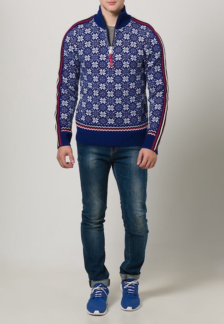 Homme portant un pull bleu à motif avec une fermeture éclair mi-longue, un jean bleu et des baskets bleues, debout devant un fond clair uni.