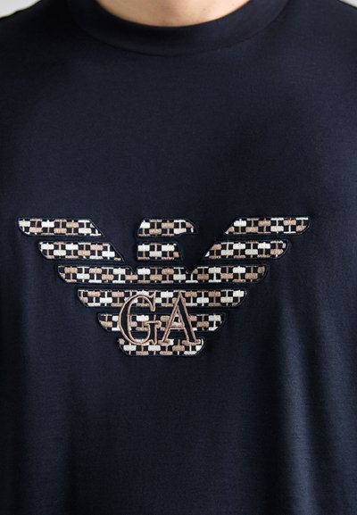 Must musta T-särk, millel on tekstuuriga tiivaga logo pruunis, kreemis ja mustas värvitoonis, mille keskel on broderiseeritud "GA".
