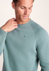 Light teal crewneck sweatshirt in a soft, smooth fabric. Features ribbed cuffs and a small logo on the chest. Slim fit and minimalist design.