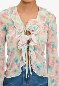 Blouse fleurie avec bords à volants, fermeture devant nouée, couleurs pastel (rose, bleu, vert) sur tissu léger et transparent. Comprend un détail de rose en 3D.