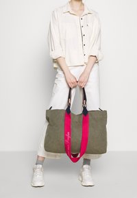 Canvas tote bag in olive green with pink straps and embroidered text. Features dark accents and a spacious design for easy carrying.