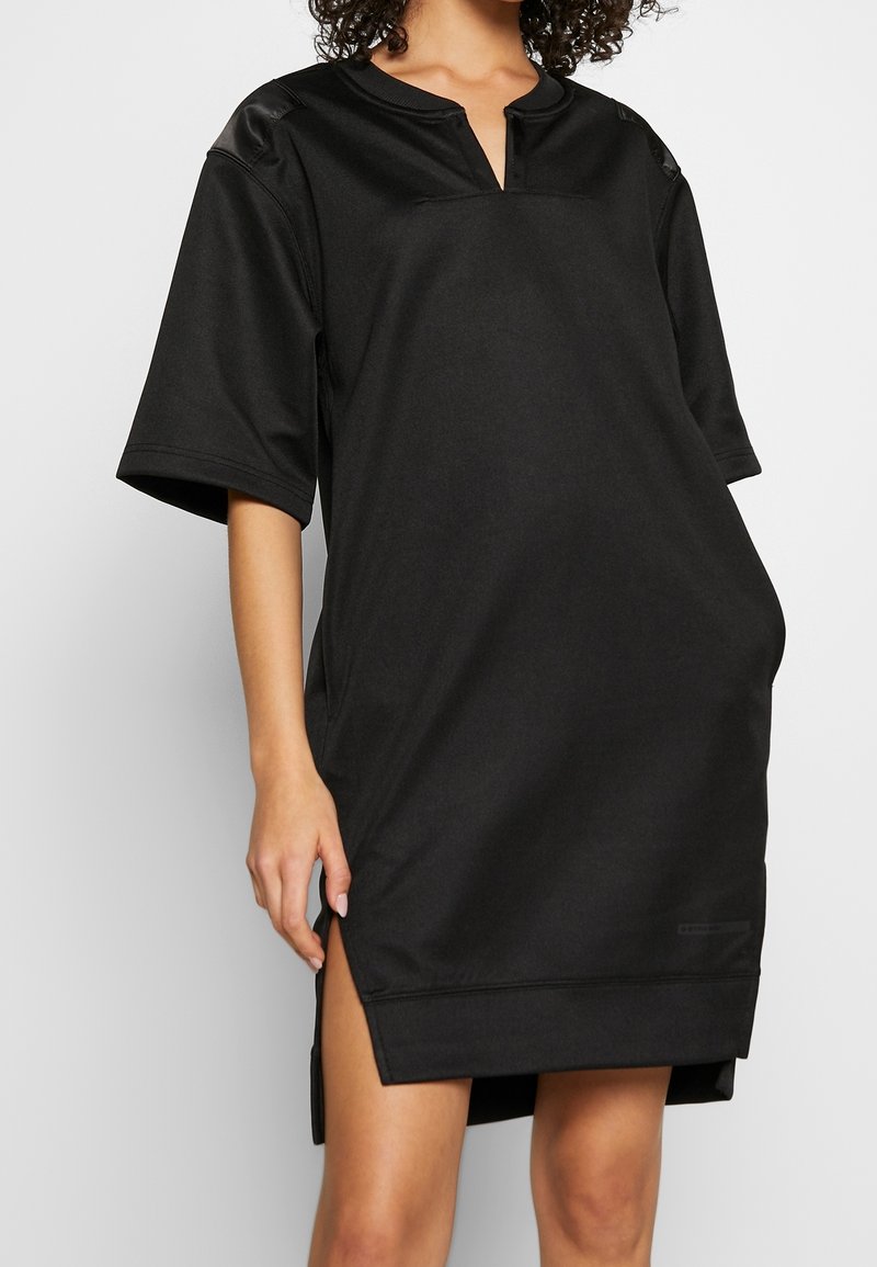 Robe noire à manches courtes en tissu lisse, avec un col en V, des épaules tombantes et des fentes latérales pour faciliter les mouvements.