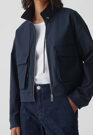 Femme portant une veste zippée bleu marine avec de grandes poches à rabat par-dessus un haut blanc et un jean bleu foncé, main dans la poche, sur un fond uni.