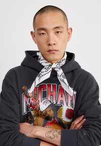 Felpa nera con una stampa grafica di un giocatore di basket, abbinata a una sciarpa bandana bianca con motivi neri. Mano con un tatuaggio visibile.