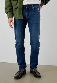 Jeans blu scuro con vestibilità slim, realizzati in denim, con cinque tasche, lievi sfumature e dettagli di cuciture marroni. Abbinati a scarpe nere.