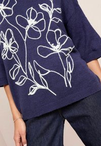 Maglione blu in maglia con ricami floreali bianchi, maniche corte, vestibilità rilassata, orlo a costine, abbinato a jeans denim scuri.