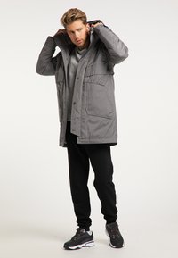 Grå isoleret parka med hætte, lynlås og knaplukning samt frontlommer. Båret over en grå skjorte og parret med sorte bukser og sneakers.