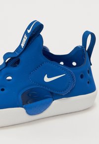 Blå sko med et teksturert syntetisk ytre, med perforeringer, en hvit Nike-logo, og en polstret såle med blå aksent.