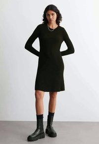 Vestido de malha preto com nervuras, de mangas longas e decote redondo, combinado com botas pretas altas e grossas e meias escuras.