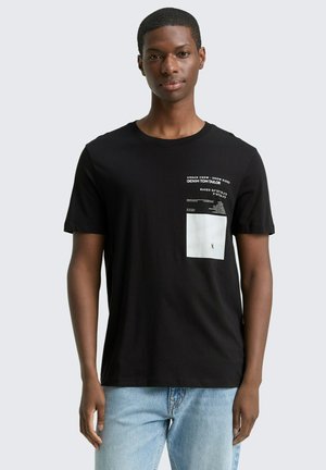 Junger Mann trägt ein schwarzes T-Shirt mit weißem Text und Grafik auf der linken Brustseite und hellblaue Jeans, steht vor einem einfarbigen Hintergrund.