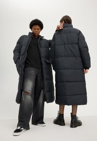 Two people wearing long black puffer coats; one faces front with hands in pockets, the other faces back wearing black boots.