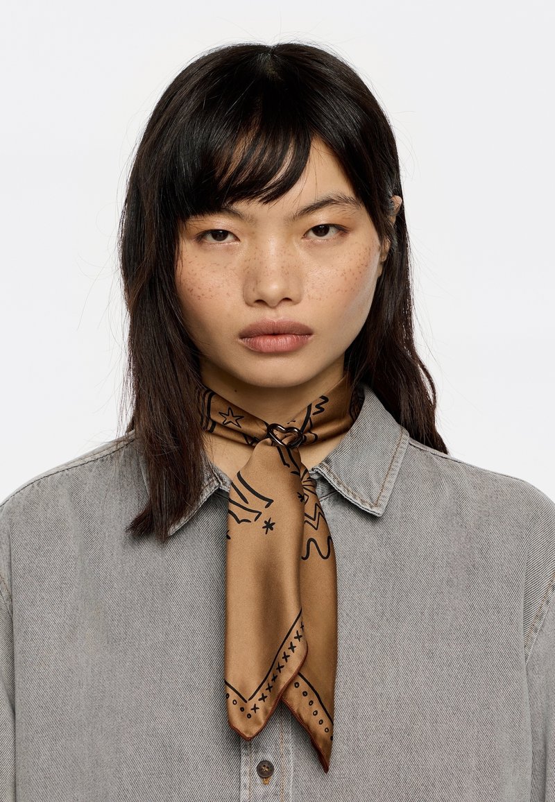 Brauner Seidenschal mit schwarzen grafischen Mustern, um den Hals gebunden. Das Model trägt ein hellgraues Jeanshemd mit Kragen.