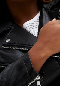 Czarna kurtka motocyklowa z sztucznej skóry z srebrnymi zamkami, z szerokim kołnierzem i fakturowanym wykończeniem. Rękaw jest lekko podwinięty.