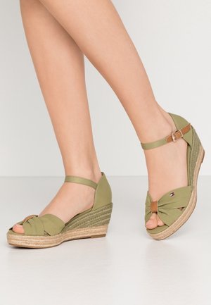 Femme portant des sandales compensées vert olive avec des brides à la cheville et des orteils ouverts, debout sur un sol clair sur fond uni.