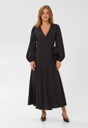 Femme portant une longue robe noire portefeuille avec manches ballon et ceinture à nouer, associée à des talons pointus foncés, debout sur fond blanc.