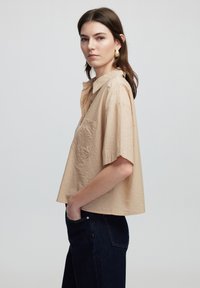 Beige cropped shirt with short sleeves, button-down front, and a pocket featuring a subtle textured pattern. Worn with dark denim pants.