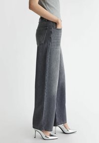 Grijze denim jeans met wijde pijpen en hoge taille, voor- en achterzakken, en een nette zoom. Gedragen met metallic zilveren high heels.