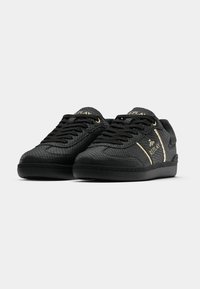 Black sneakers with textured material, gold accents, and laces, featuring a low-top design and rubber outsole. Notable logo on the side.