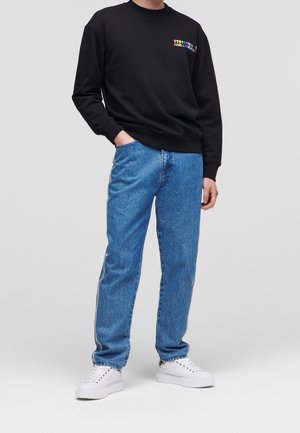 Person trägt einen schwarzen Pullover mit einem bunten Logo, blaue Jeans mit seitlichen Streifen, weiße Turnschuhe und steht mit einer Hand in der Tasche.