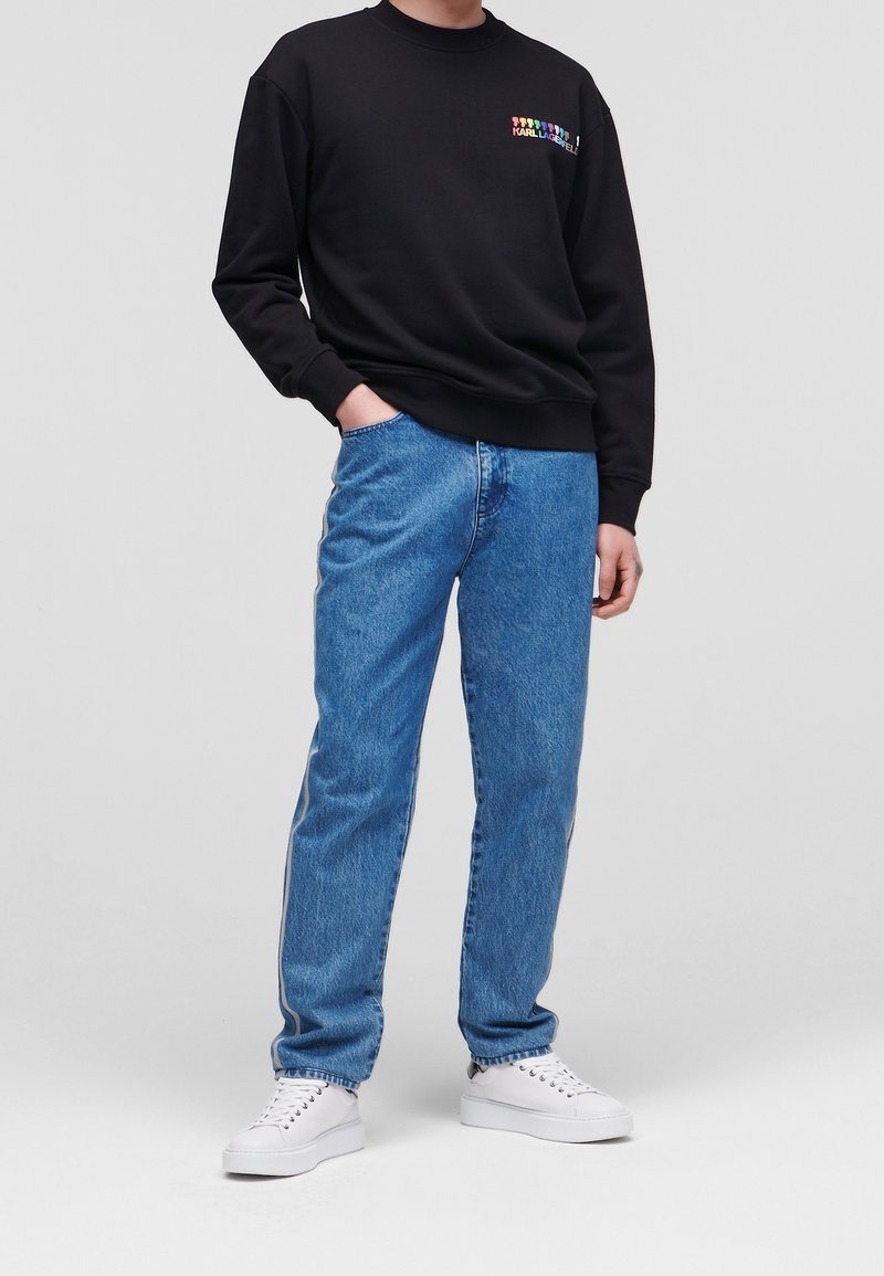 Personne portant un sweat-shirt noir avec un logo coloré, un jean bleu avec des bandes latérales, des baskets blanches, debout avec une main dans la poche.